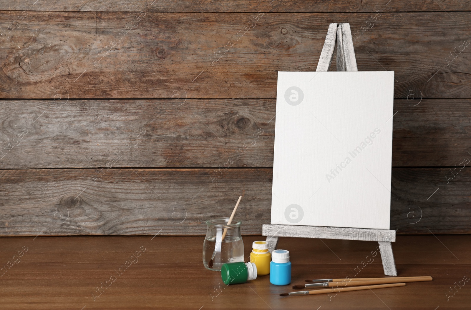 Photo of Easel with blank canvas board and painting tools for children on table near wooden wall. Space for text