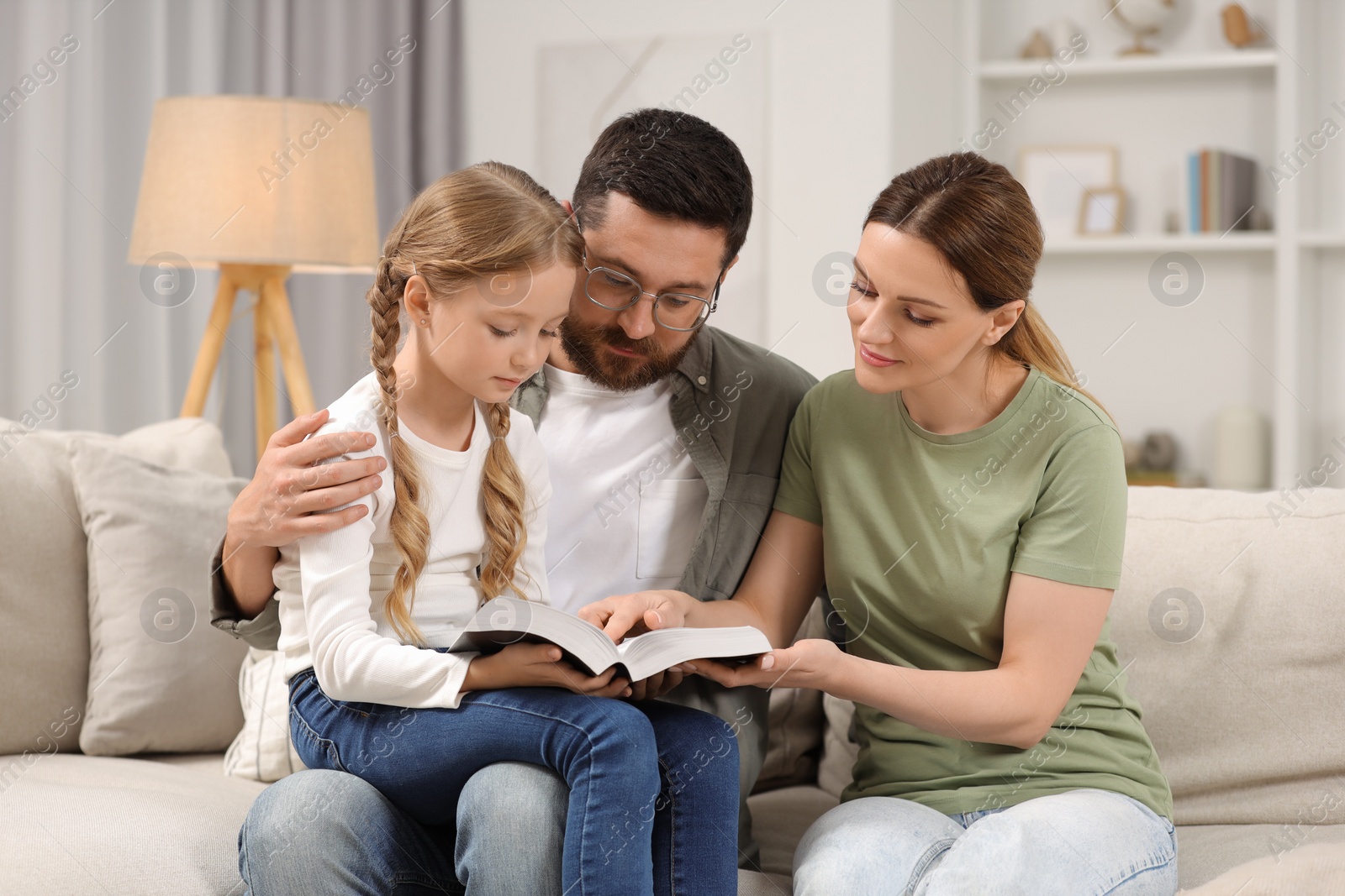 Photo of Girl and her godparents reading Bible together on sofa at home