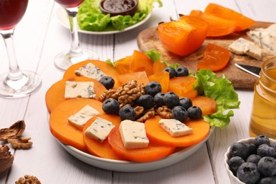Photo of Delicious persimmon with blue cheese, blueberries, lettuce and nuts on white wooden table, closeup