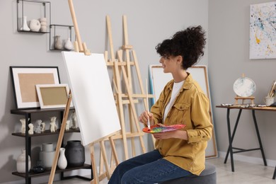 Photo of Young woman mixing paints on palette with brush near easel in studio