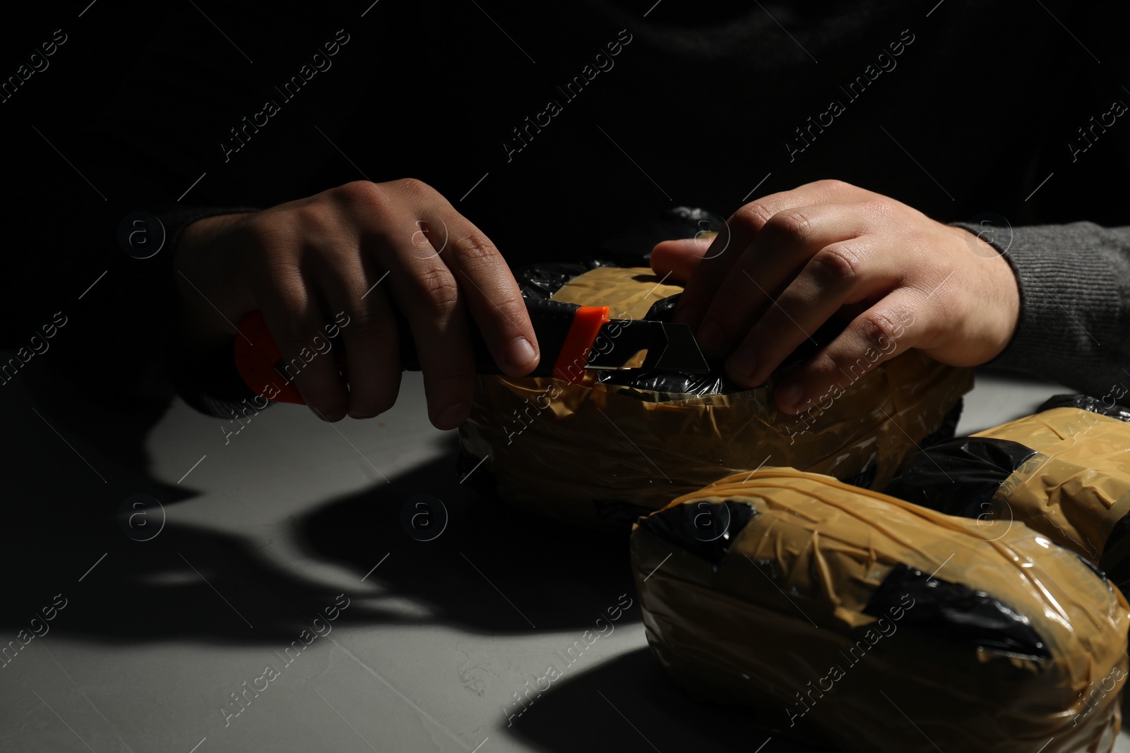 Photo of Smuggling and drug trafficking. Man opening package of narcotics with box cutter in darkness, closeup