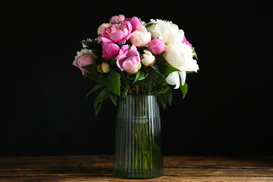 Bouquet of beautiful peonies in vase on wooden table against black background