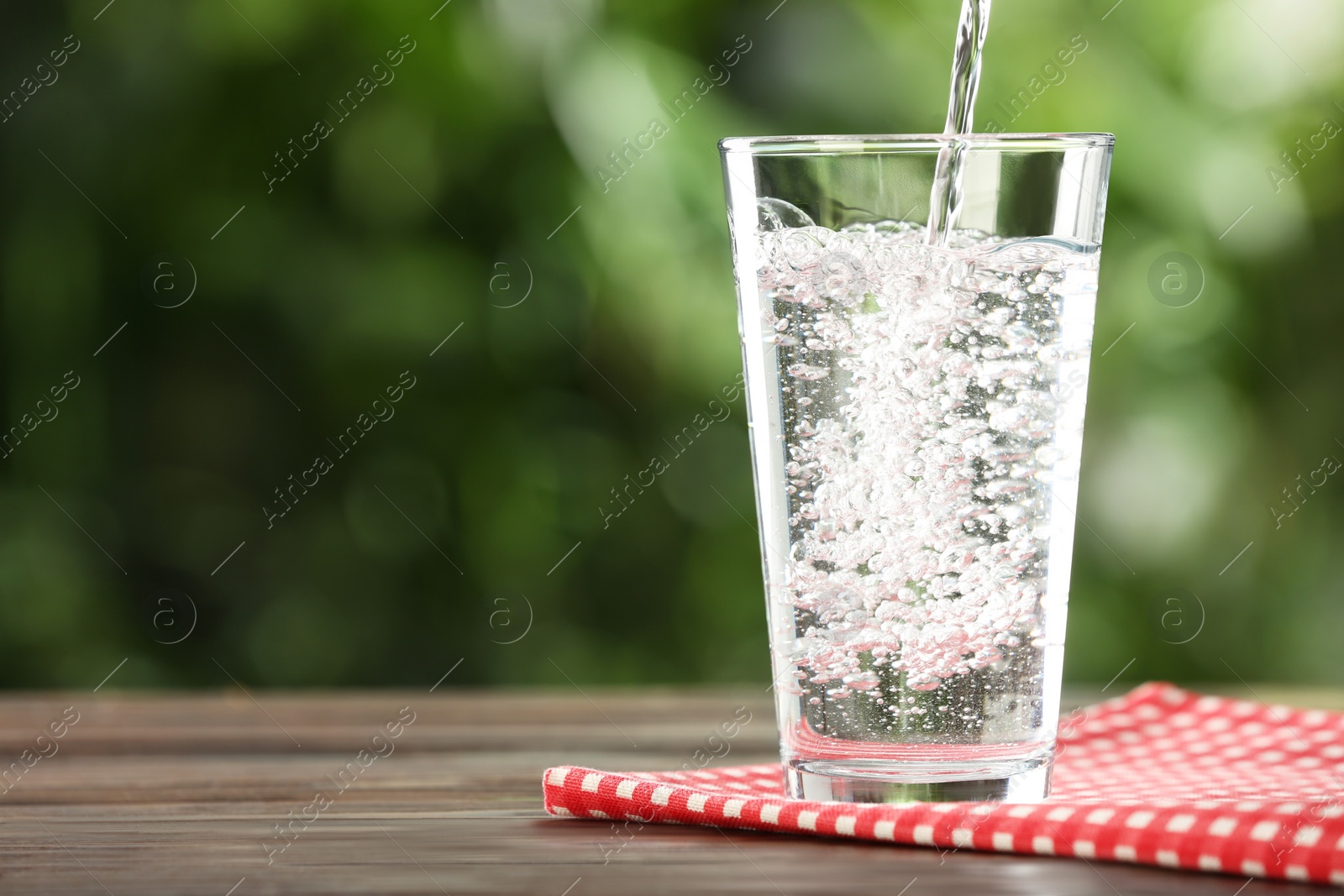 Photo of Pouring water into glass on wooden table outdoors, space for text