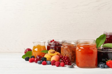 Photo of Jars with different jams and fresh fruits on white wooden table. Space for text