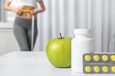 Photo of Weight loss pills with apple on table and woman measuring waist indoors. Space for text