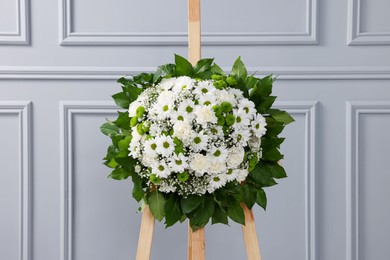 Funeral wreath of flowers on wooden stand near light grey wall