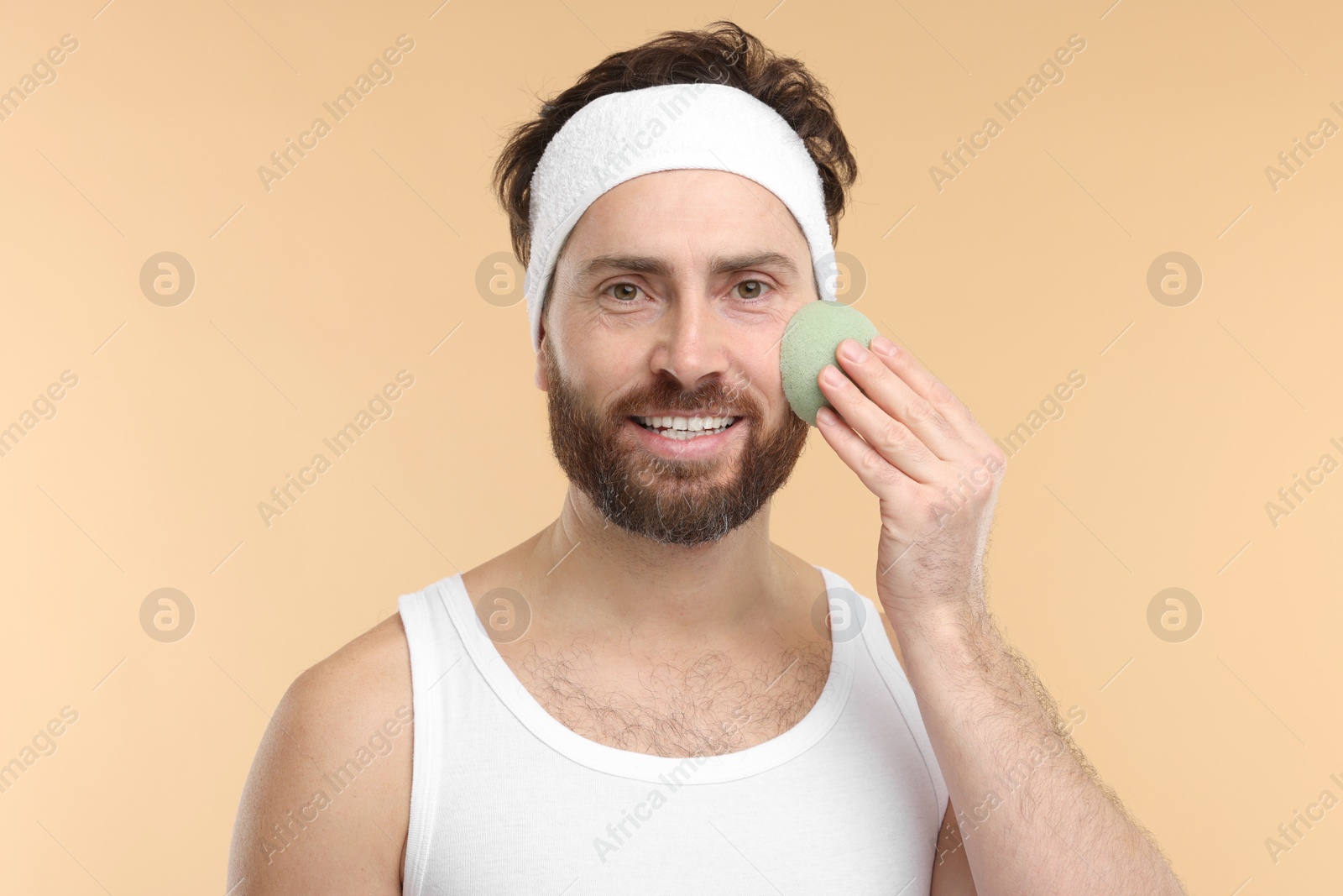 Photo of Man with headband washing his face using sponge on beige background