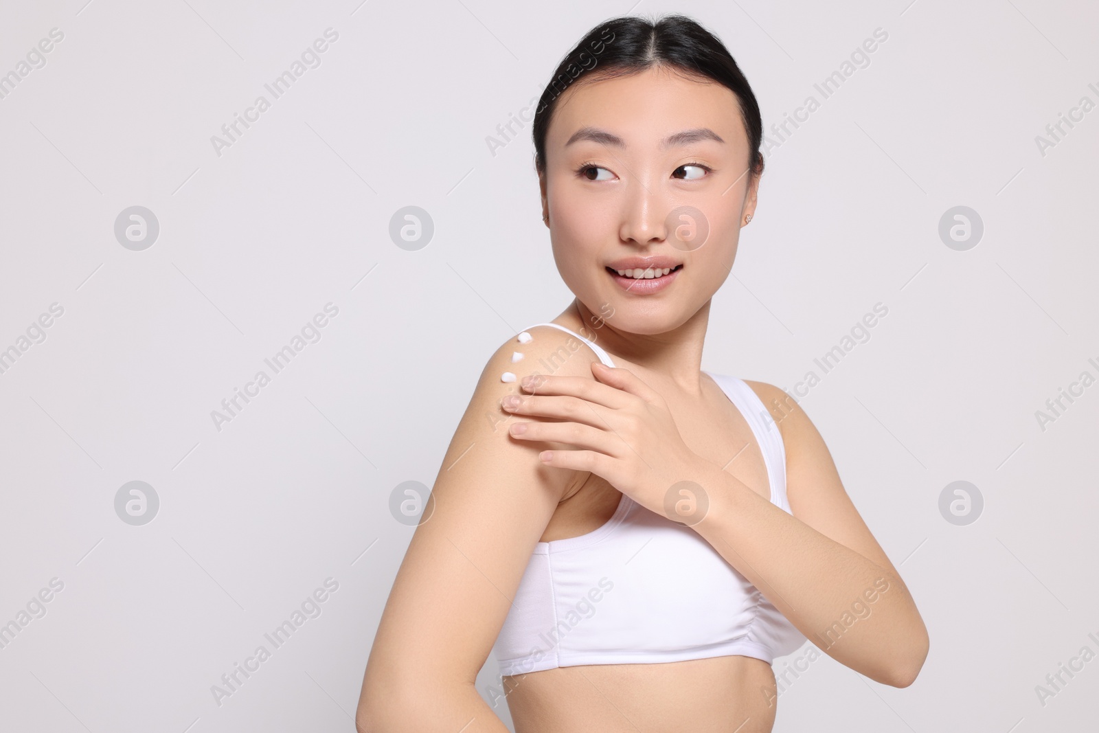 Photo of Beautiful young Asian woman applying body cream onto shoulder on light grey background, space for text