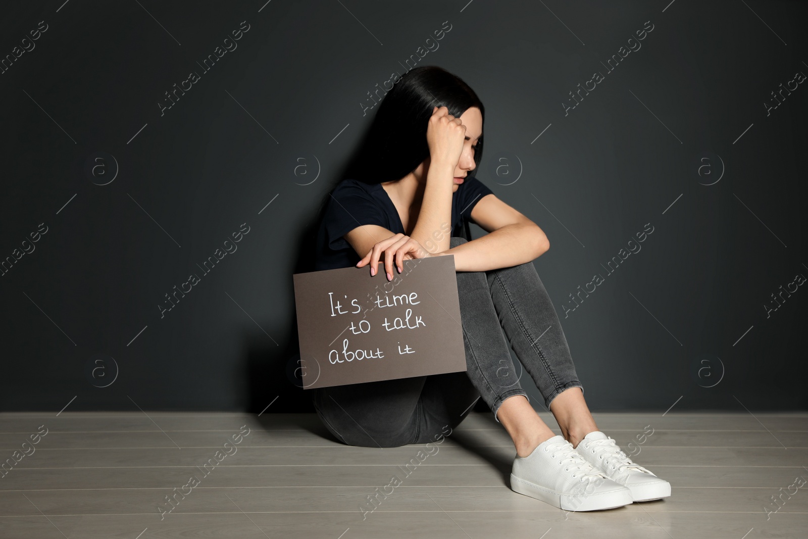 Photo of Young woman holding card with words IT'S TIME TO TALK ABOUT IT while sitting near grey wall