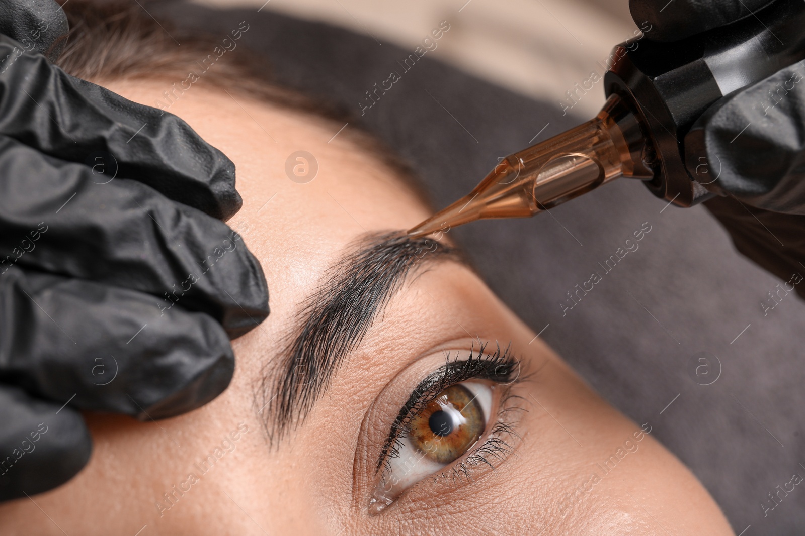 Photo of Young woman undergoing procedure of permanent eyebrow makeup in tattoo salon, closeup