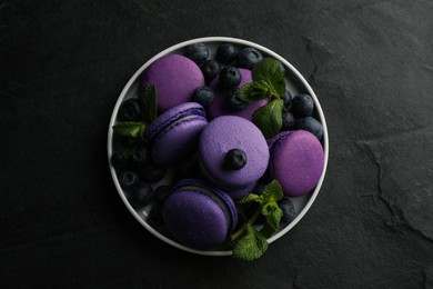 Photo of Delicious macarons, mint and blueberries on black table, top view