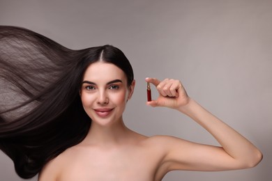 Beautiful young woman with long healthy hair holding ampoule on grey background