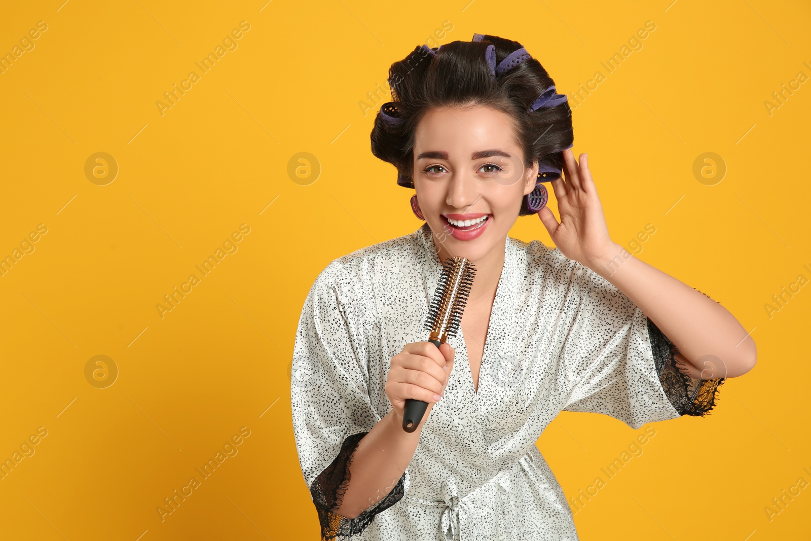 Photo of Happy young woman in silk bathrobe with hair curlers singing into hairbrush on orange background, space for text