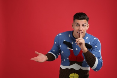 Photo of Man in Christmas sweater showing silence gesture on red background, space for text