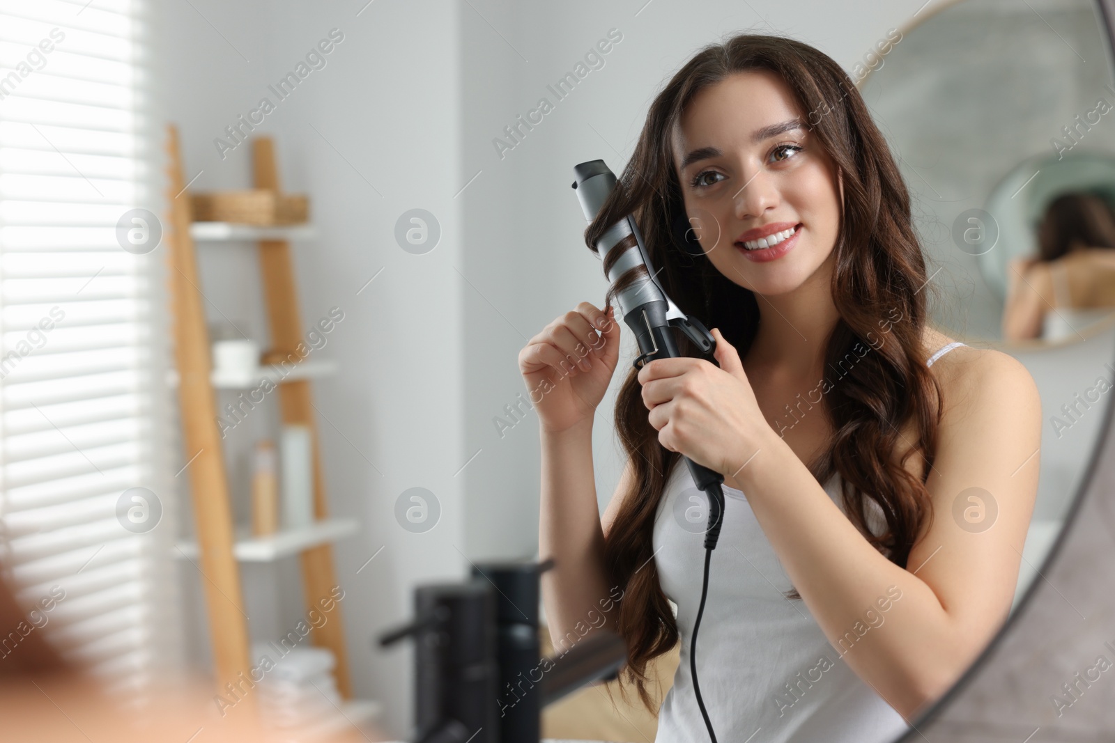 Photo of Smiling woman using curling hair iron near mirror in bathroom