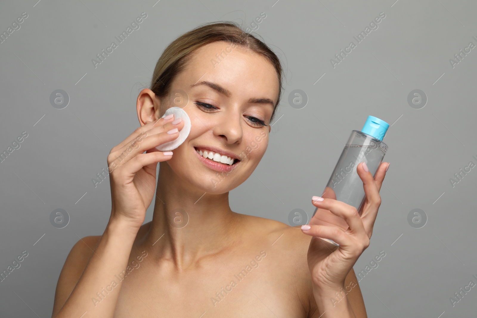 Photo of Smiling woman removing makeup with cotton pad and holding bottle on grey background