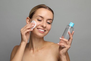 Photo of Smiling woman removing makeup with cotton pad and holding bottle on grey background