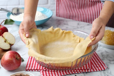 Woman putting dough for apple pie into baking dish at white marble table, closeup
