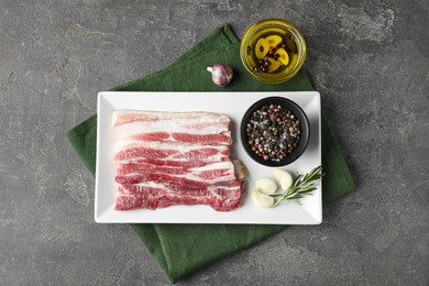 Photo of Pieces of raw pork belly, oil, peppercorns, rosemary and garlic on grey textured table, flat lay