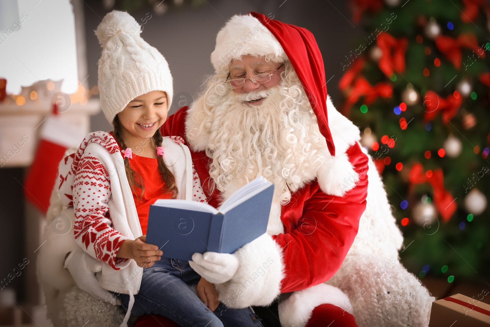 Photo of Little girl reading book while sitting on authentic Santa Claus' lap indoors