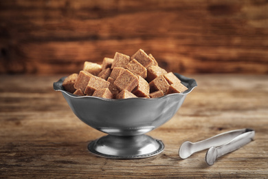 Photo of Brown sugar cubes in metal bowl and tongs on wooden table
