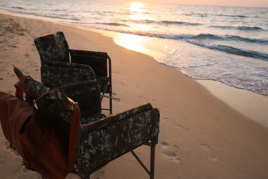 Camping chairs and backpack on sandy beach near sea, space for text