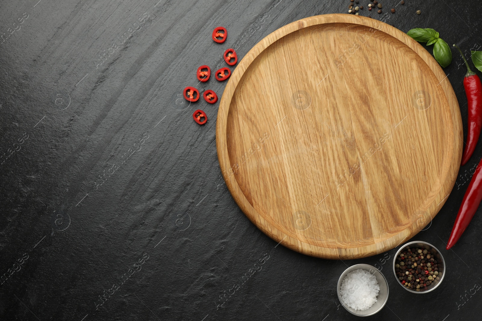 Photo of Cutting board, salt, spices, basil and chili peppers on black textured table, flat lay. Space for text