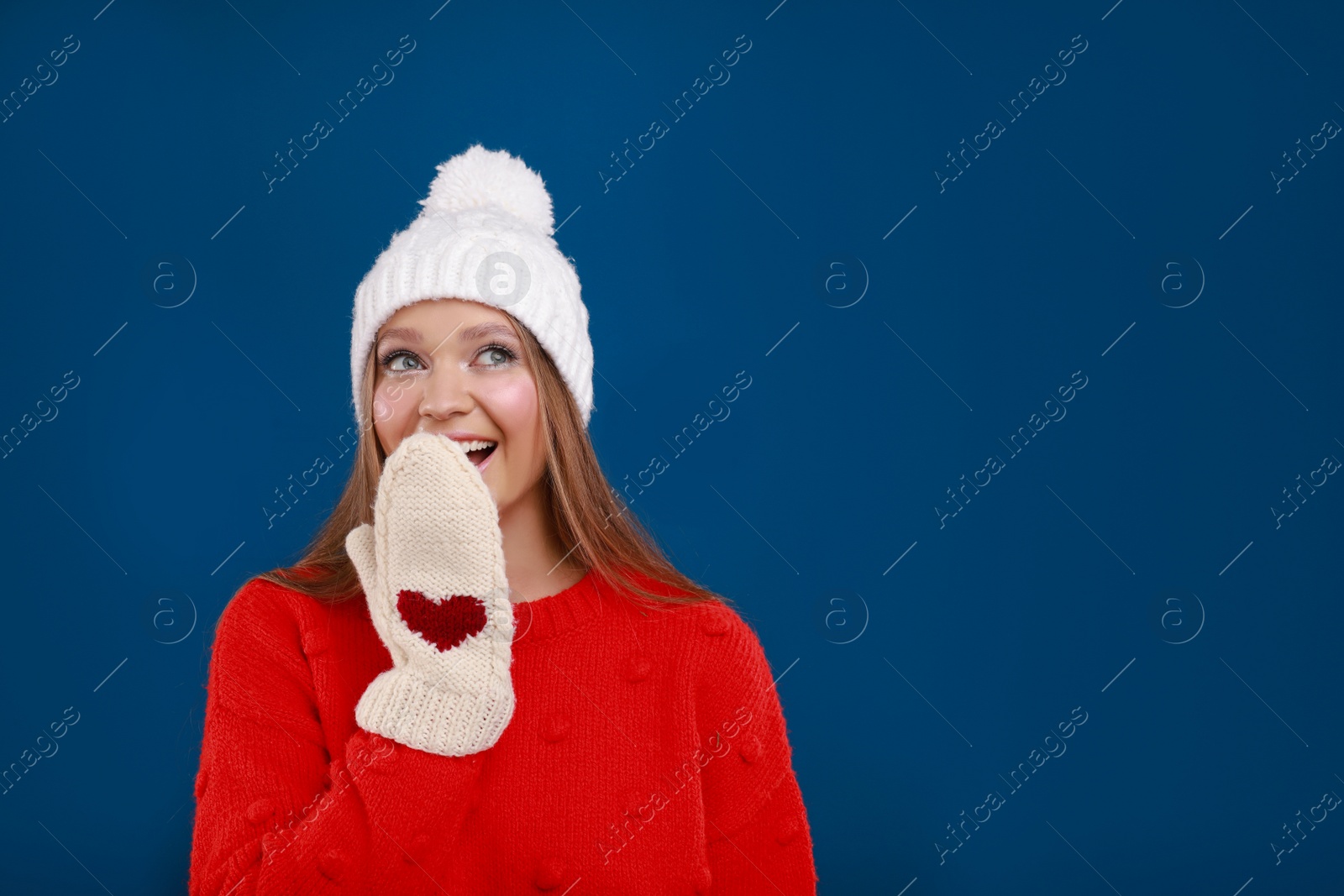 Photo of Emotional young woman in warm sweater, mittens and hat on blue background, space for text. Winter season