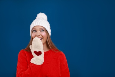 Photo of Emotional young woman in warm sweater, mittens and hat on blue background, space for text. Winter season