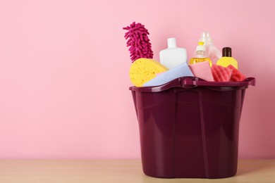 Bucket with different cleaning supplies on wooden floor near pink wall. Space for text