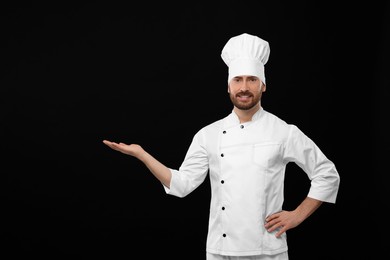 Smiling mature chef showing something on black background, space for text
