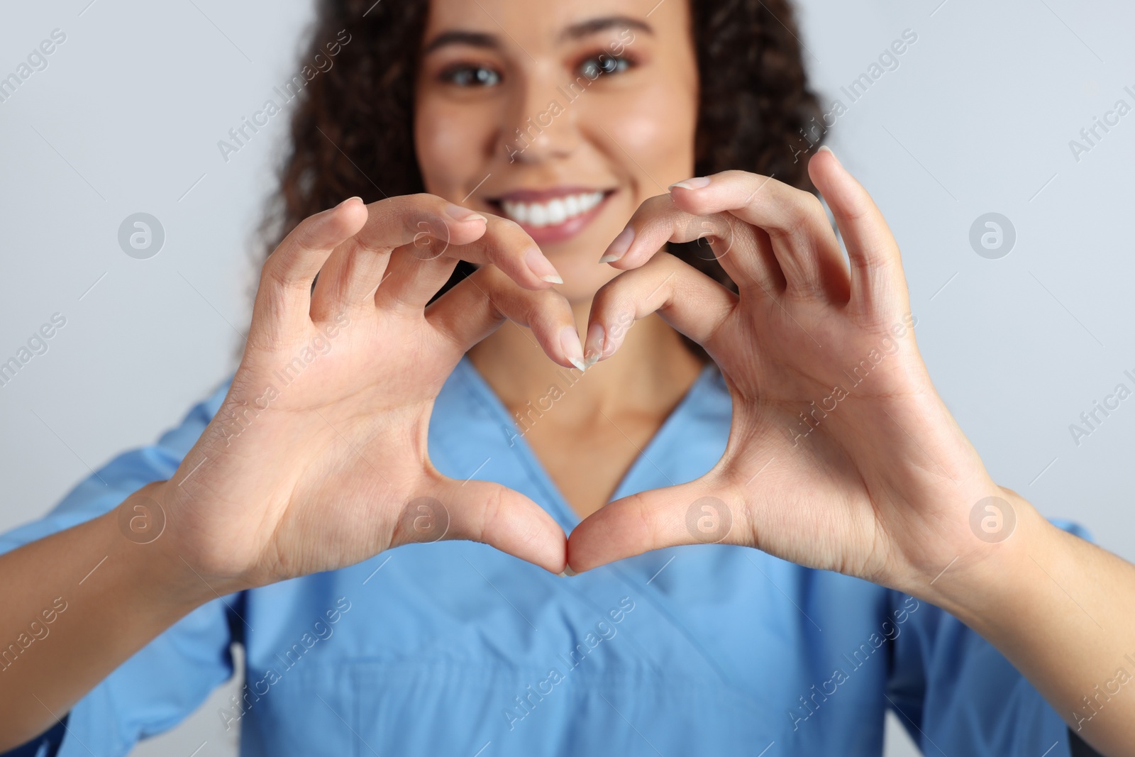 Photo of Happy young African-American doctor making heart with hands on light grey background, closeup