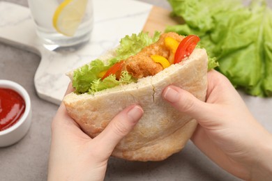 Woman holding delicious pita sandwich with fried fish, pepper, tomatoes and lettuce at light grey table, closeup