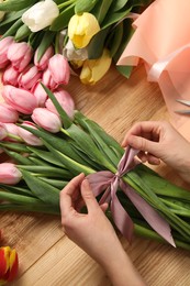 Photo of Woman making beautiful bouquet of fresh tulips and ribbon at wooden table, top view