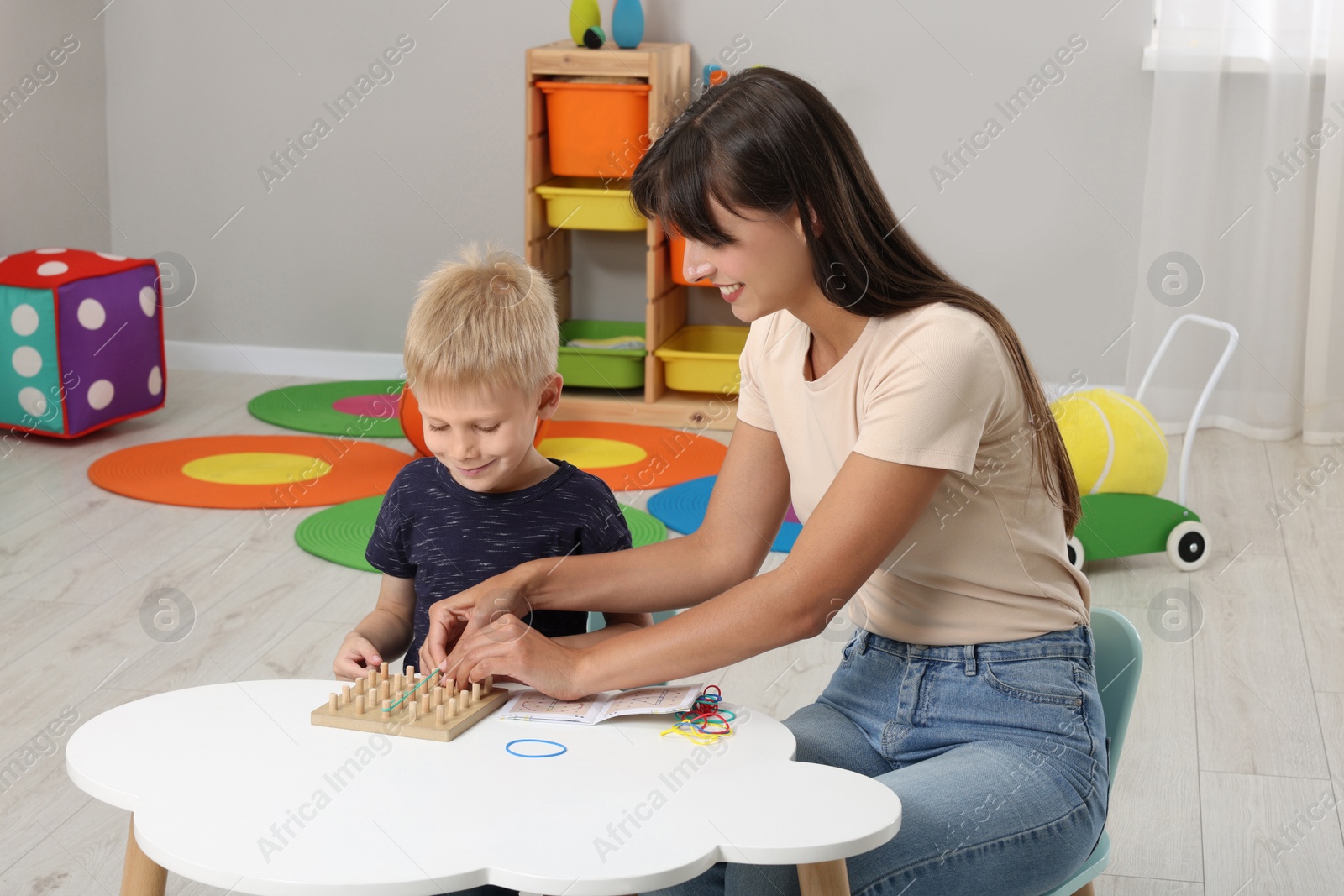 Photo of Motor skills development. Happy mother helping her son to play with geoboard and rubber bands at white table in room