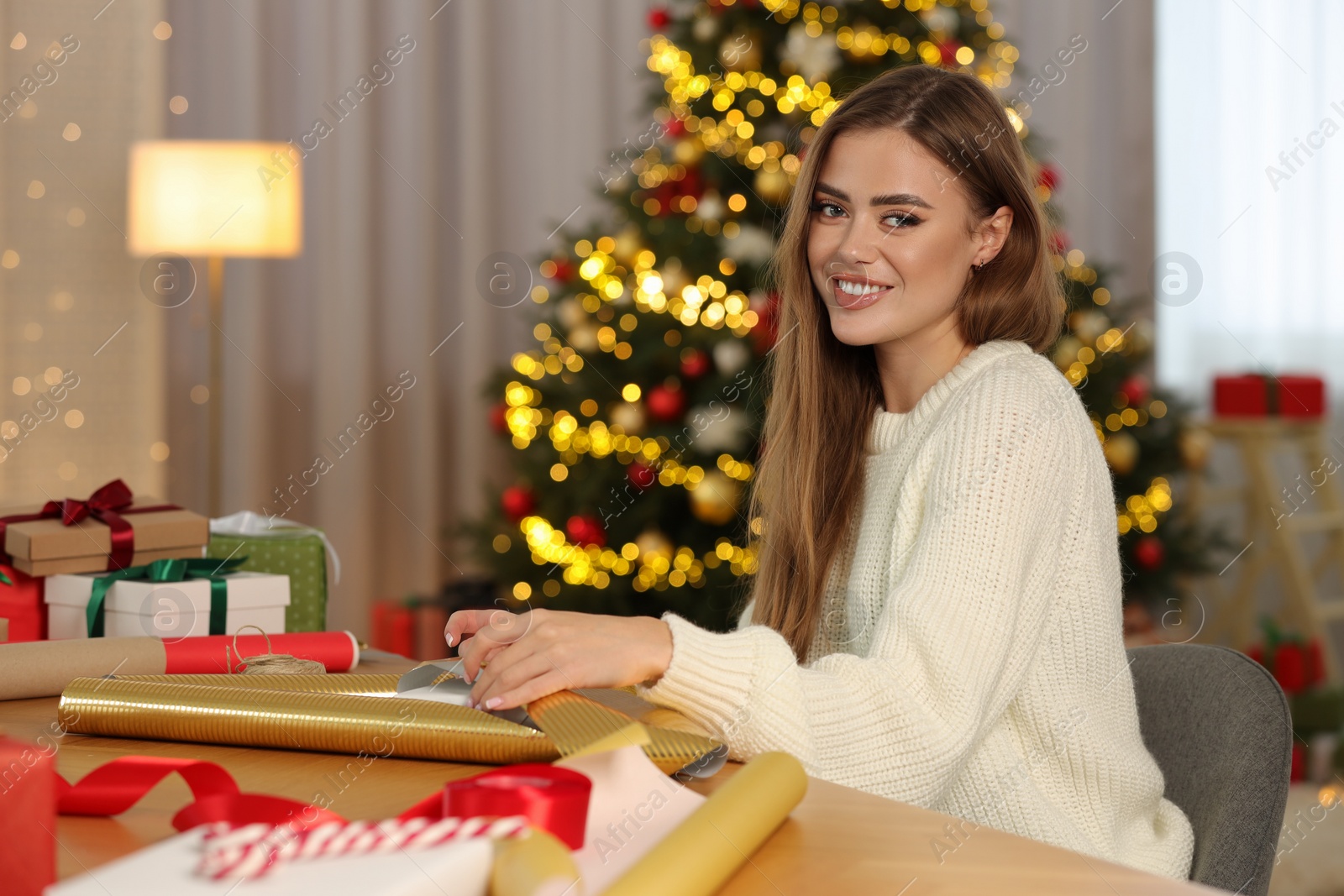 Photo of Beautiful young woman with wrapping paper at table in room. Decorating Christmas gift