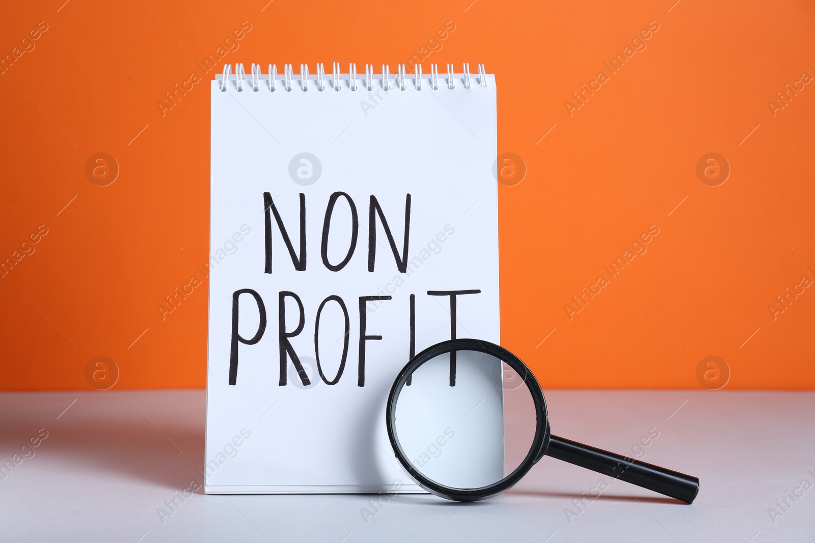 Photo of Notebook with phrase Non Profit and magnifying glass on white table