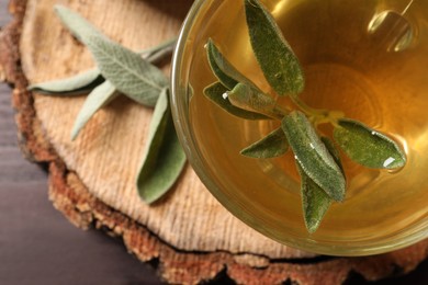 Photo of Cup of aromatic sage tea with fresh leaves on wooden stump, closeup