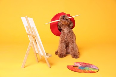 Photo of Cute Maltipoo in red beret holding brush near easel with canvas and palette on orange background. Dog artist