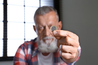 Senior man holding coin indoors, focus on hand. Space for text