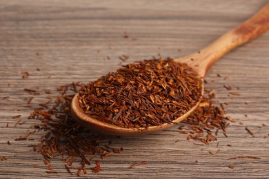 Photo of Spoon with dry rooibos leaves on wooden table, closeup