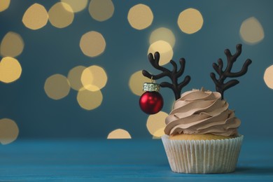 Photo of Tasty Christmas cupcake with chocolate reindeer antlers and bauble on blue wooden table against blurred festive lights, space for text