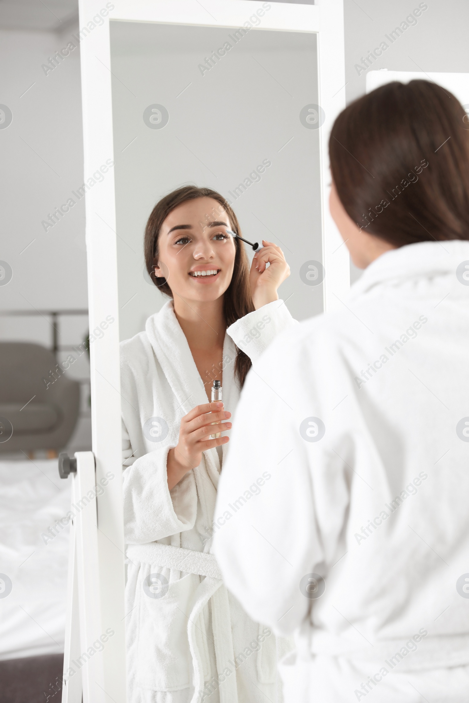 Photo of Attractive young woman applying oil onto her eyelashes near mirror indoors