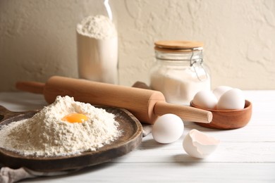 Photo of Making dough. Pile of flour with yolk, rolling pin and eggs on white wooden table, closeup