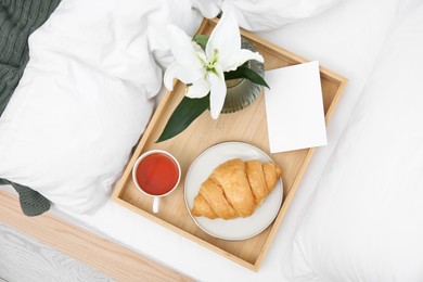 Photo of Tray with tasty croissant, cup of tea and flower on white bed, top view
