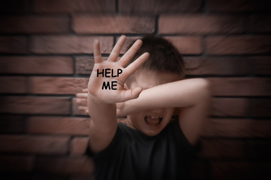 Scared little boy with text HELP ME on his hand near brick wall
