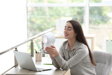 Photo of Young woman enjoying air flow from portable fan at workplace. Summer heat