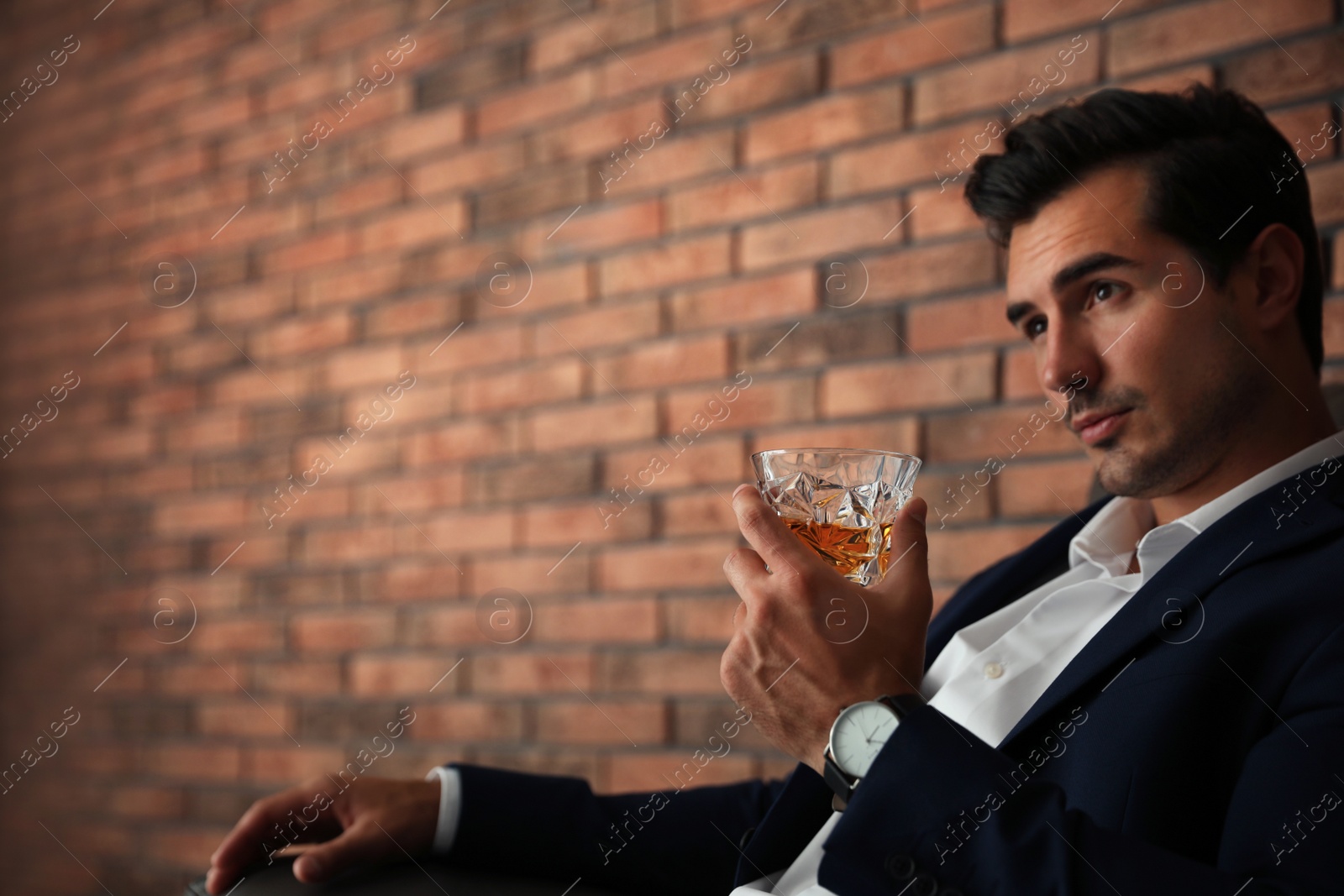 Photo of Young man with glass of whiskey near brick wall indoors. Space for text