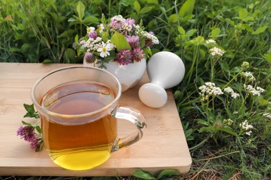 Photo of Cup of aromatic herbal tea, pestle and ceramic mortar with different wildflowers on green grass outdoors. Space for text
