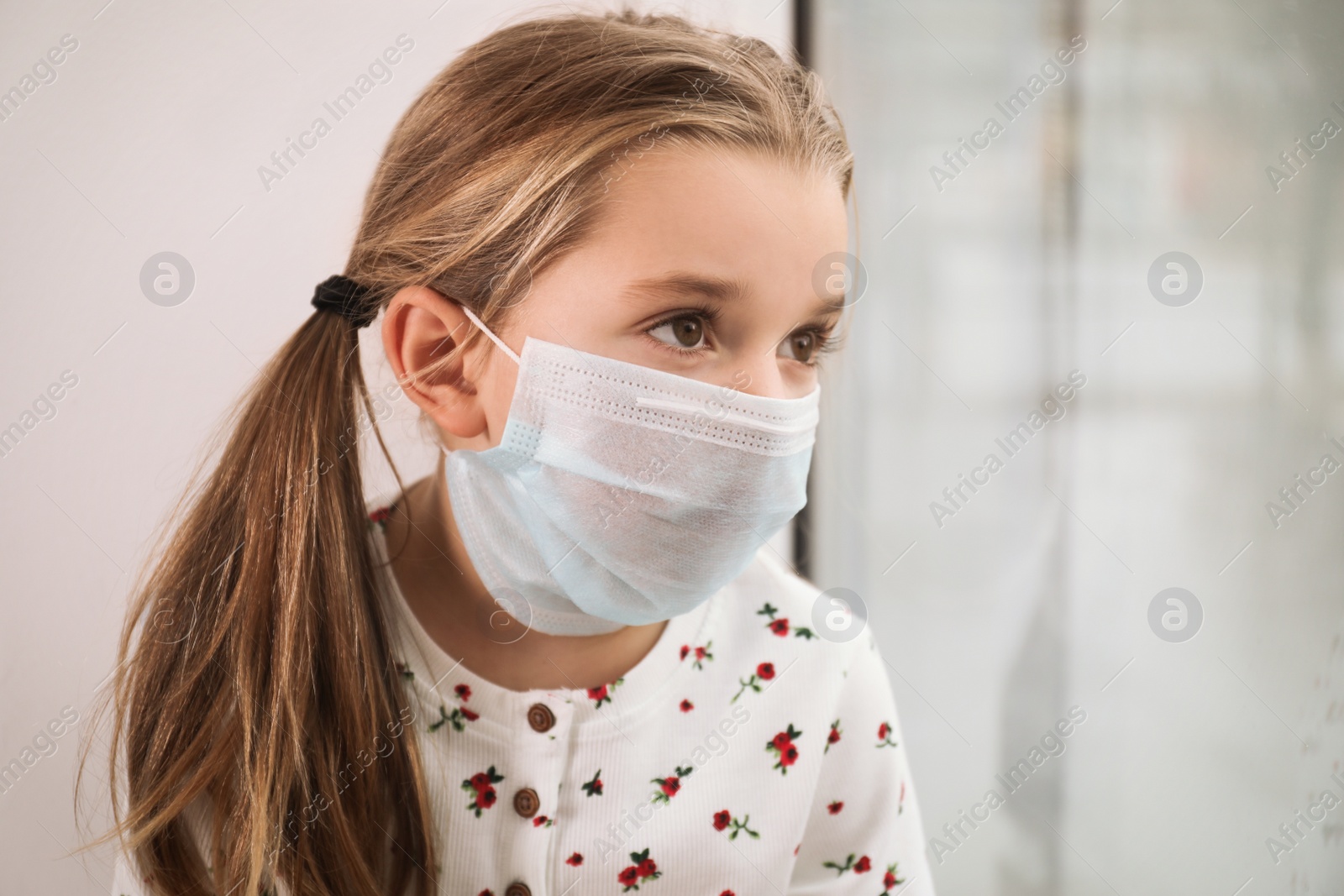 Photo of Sad little girl in protective mask looking out of window indoors. Staying at home during coronavirus pandemic